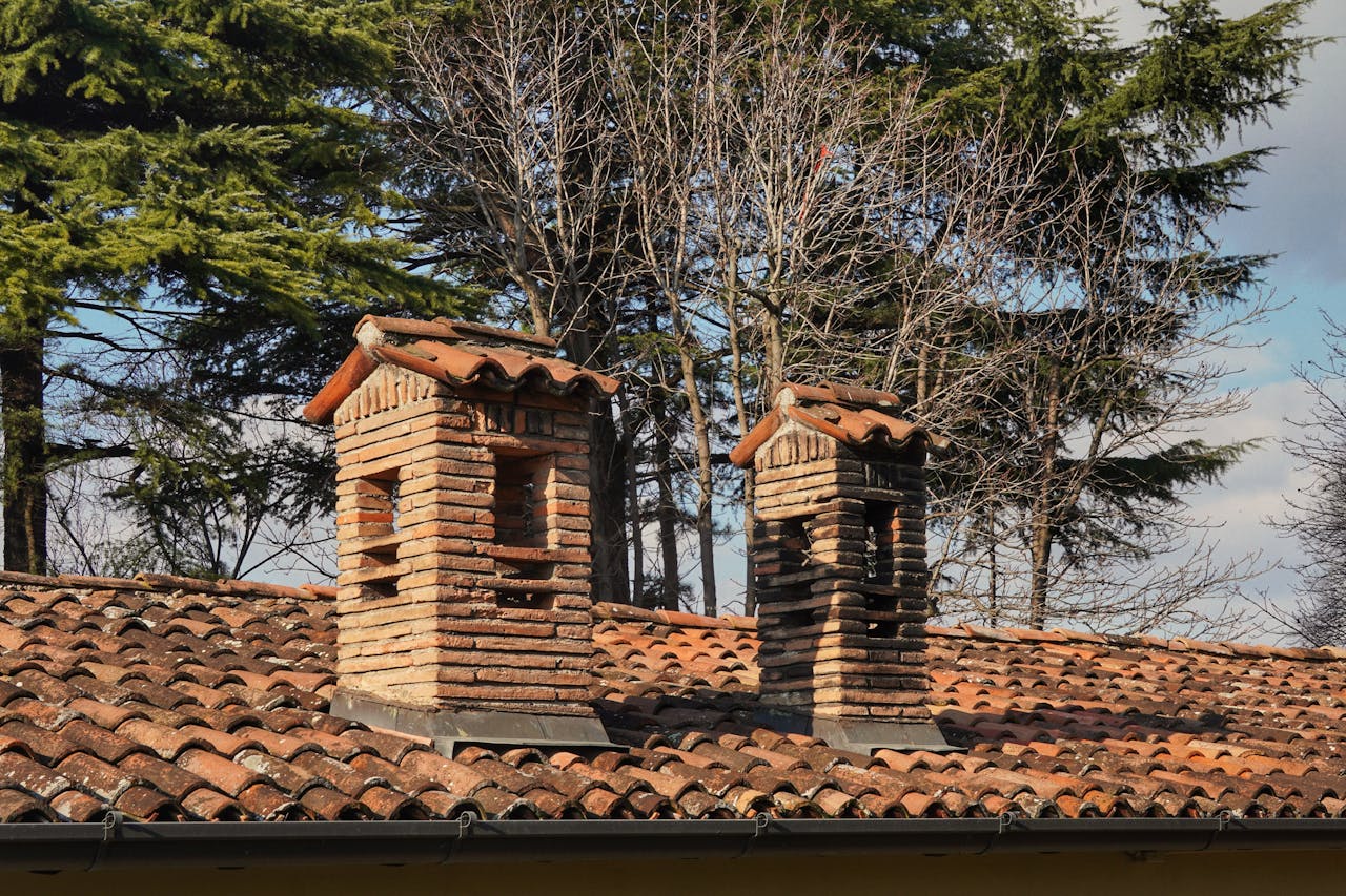 Two rustic brick chimneys on a red-tiled roof with a winter forest backdrop.