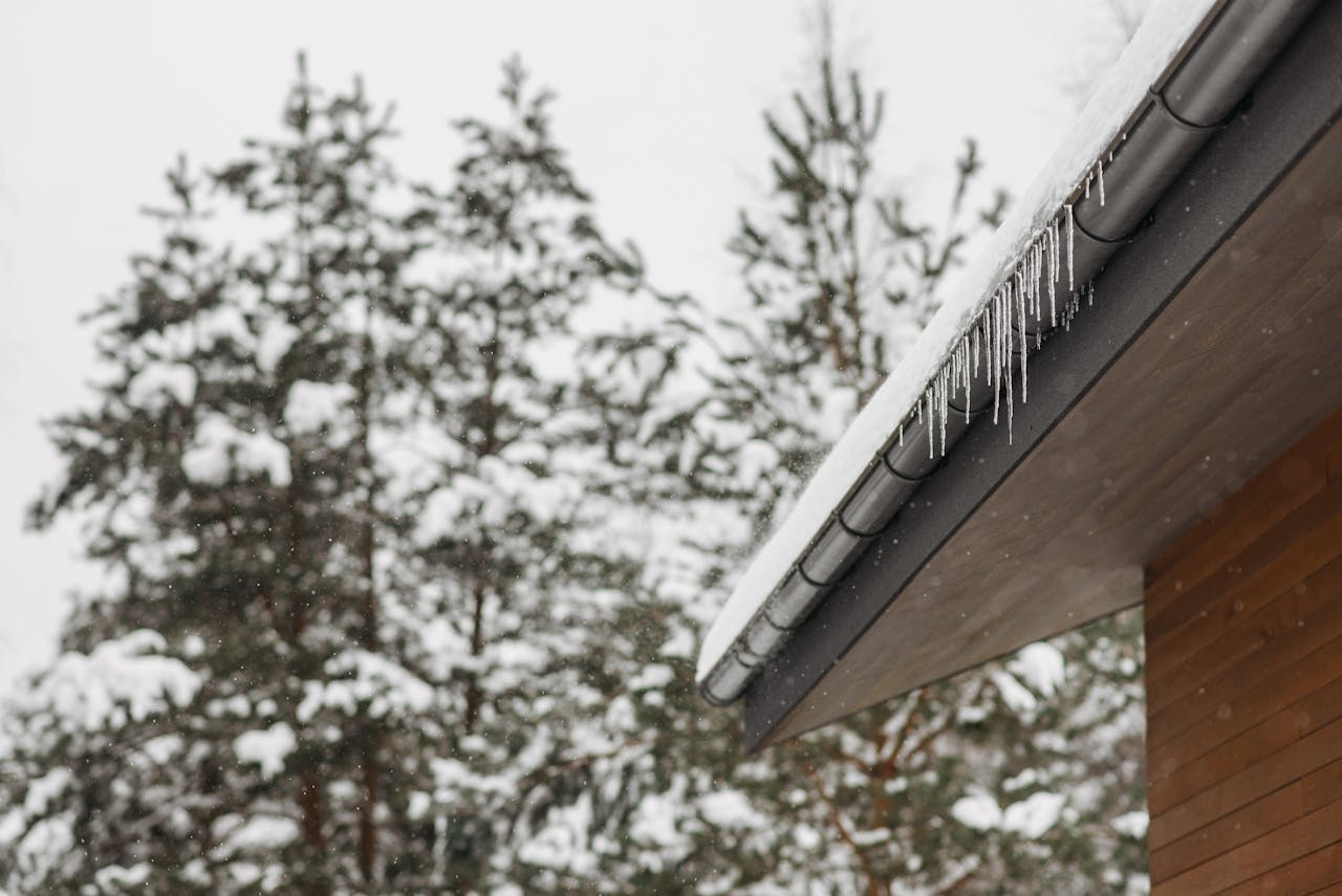 services-05 Stunning winter scene of icicles hanging from a snow-covered roof with snowy pines in the background.